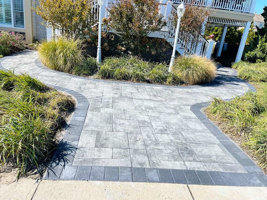 Grey walkway with dark grey bordering, surrounded by grass and plants.